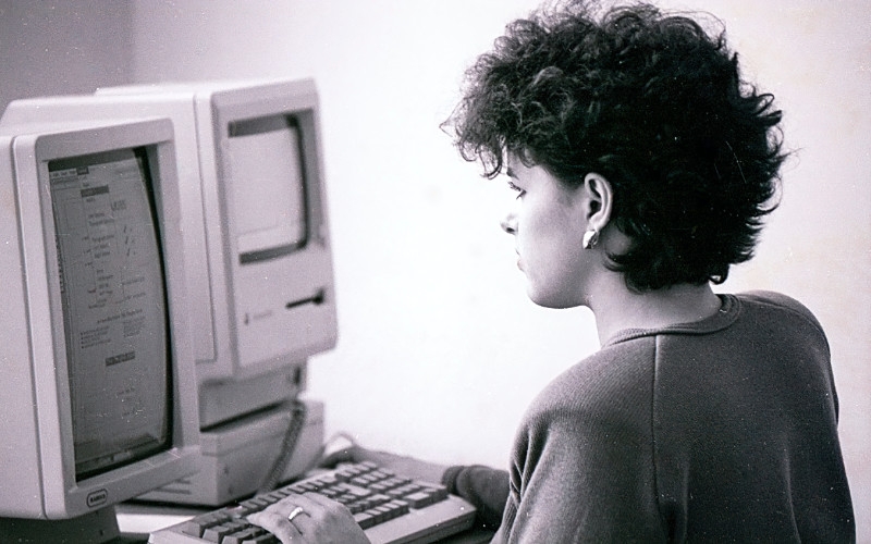 a black and white picture of a woman staring at an old computer while using the keyboard to type