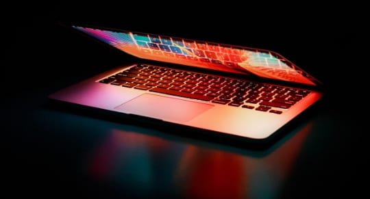 a glowing laptop computer sits in a dark room, illuminating its screen and keyboard