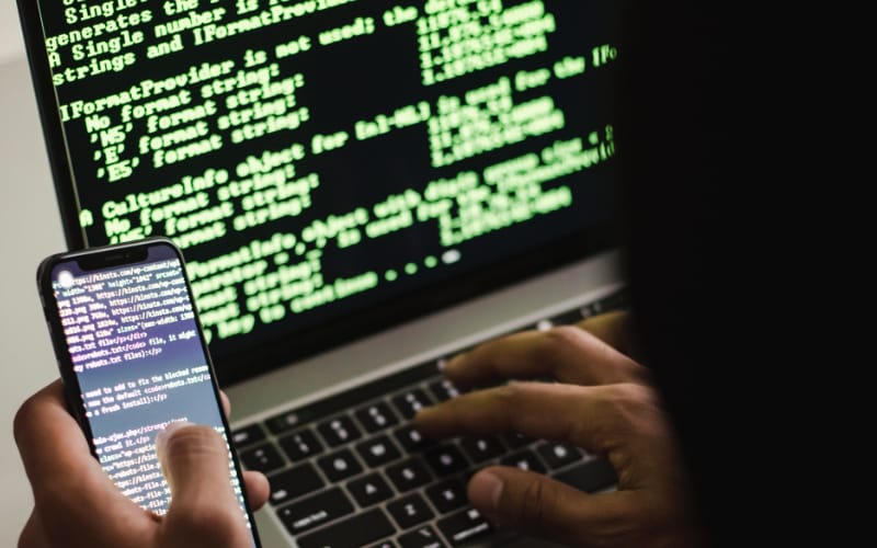 a person typing on a laptop displaying green code on a black screen, holding a smartphone showing similar texts