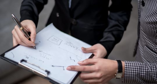 two professionals in business attire holding a clipboard, engaged in discussion in an office setting