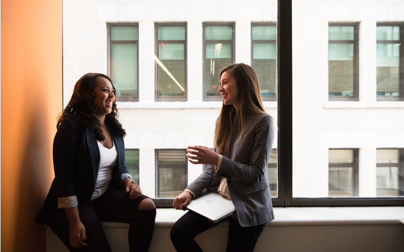 The culture needs to empower everyone to help break down barriers two women engaged in conversation while standing by an office window, with natural light illuminating the space